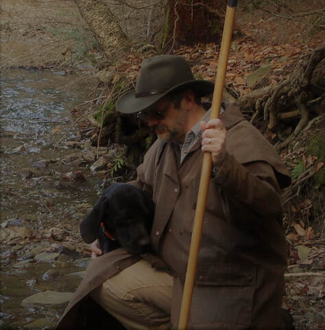 Picture of the author kneeling beside a creek with a large black dog. The author is wearing a brown drover and brown fedora. He's also holding a staff.